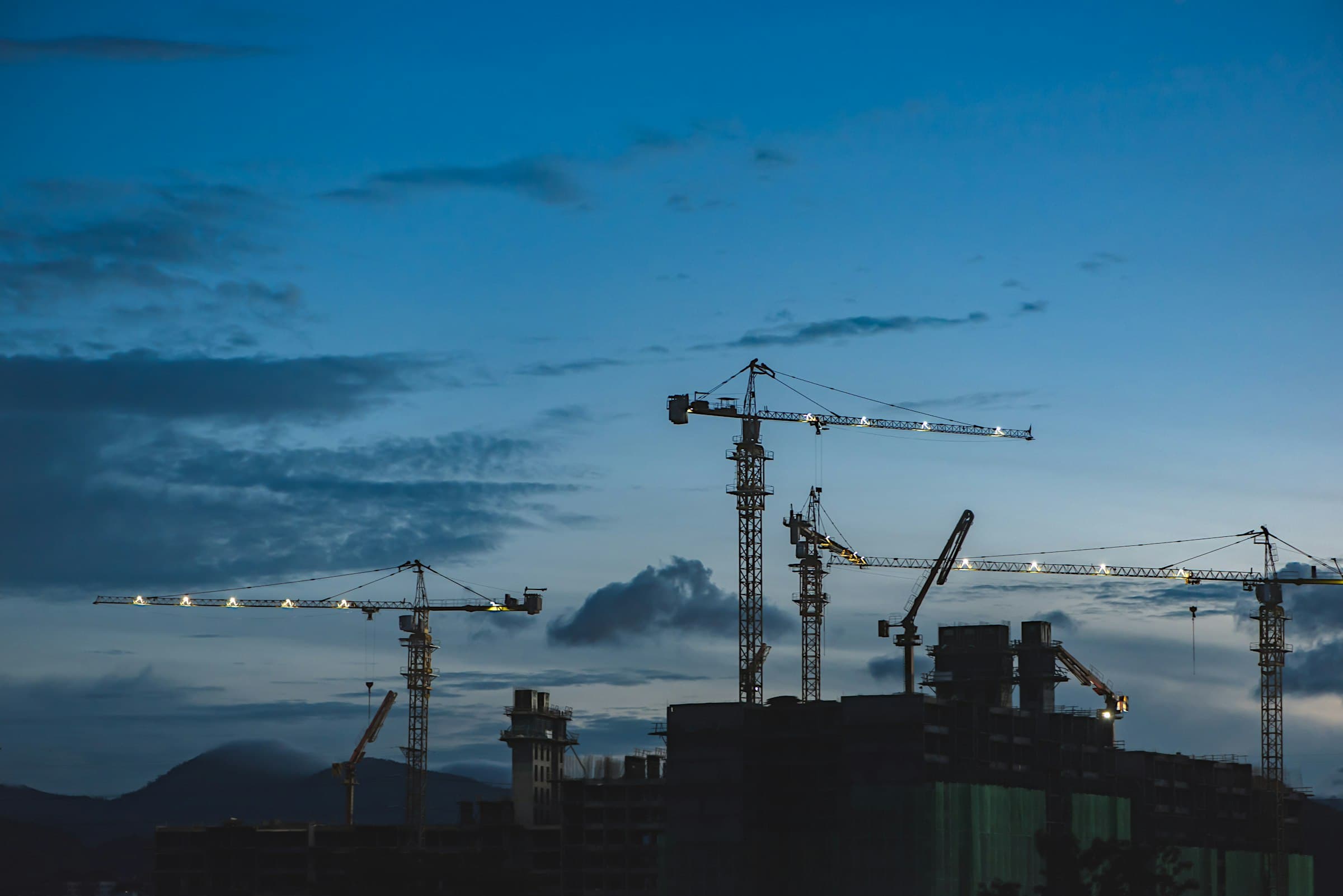 Construction cranes silhouetted against a deep blue dusk sky